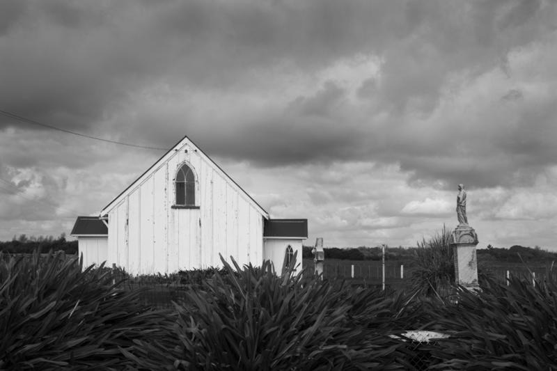 A black and white photo of a church under a cloudy sky and with a harakeke hedge in front