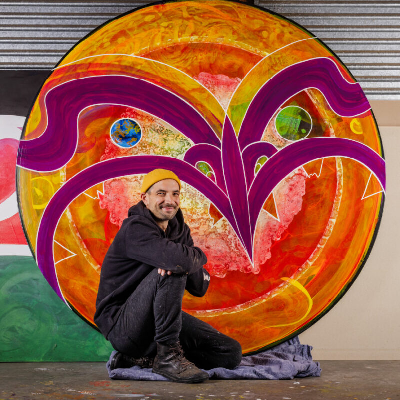 photograph of an artist sitting in front of his colourful circular artwork
